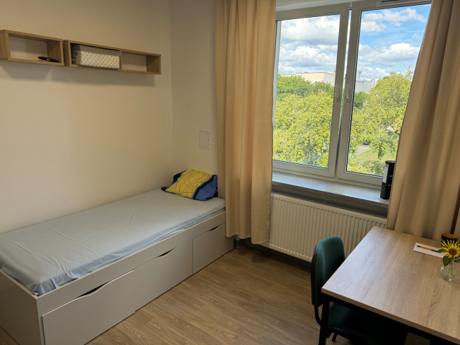 Pokój jednoosobowy w DS. Grosik. Po wejściu na wprost znajduje się duże okno z żółtymi zasłonami. Po lewej stronie widoczne jest łóżko z szufladami do przechowywania, a po prawej stronie drewniane biurko z krzesłem. Pokój jest jasny i przestronny.